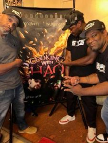 Three men pointing at a Cooking with Chaos poster displayed on an easel